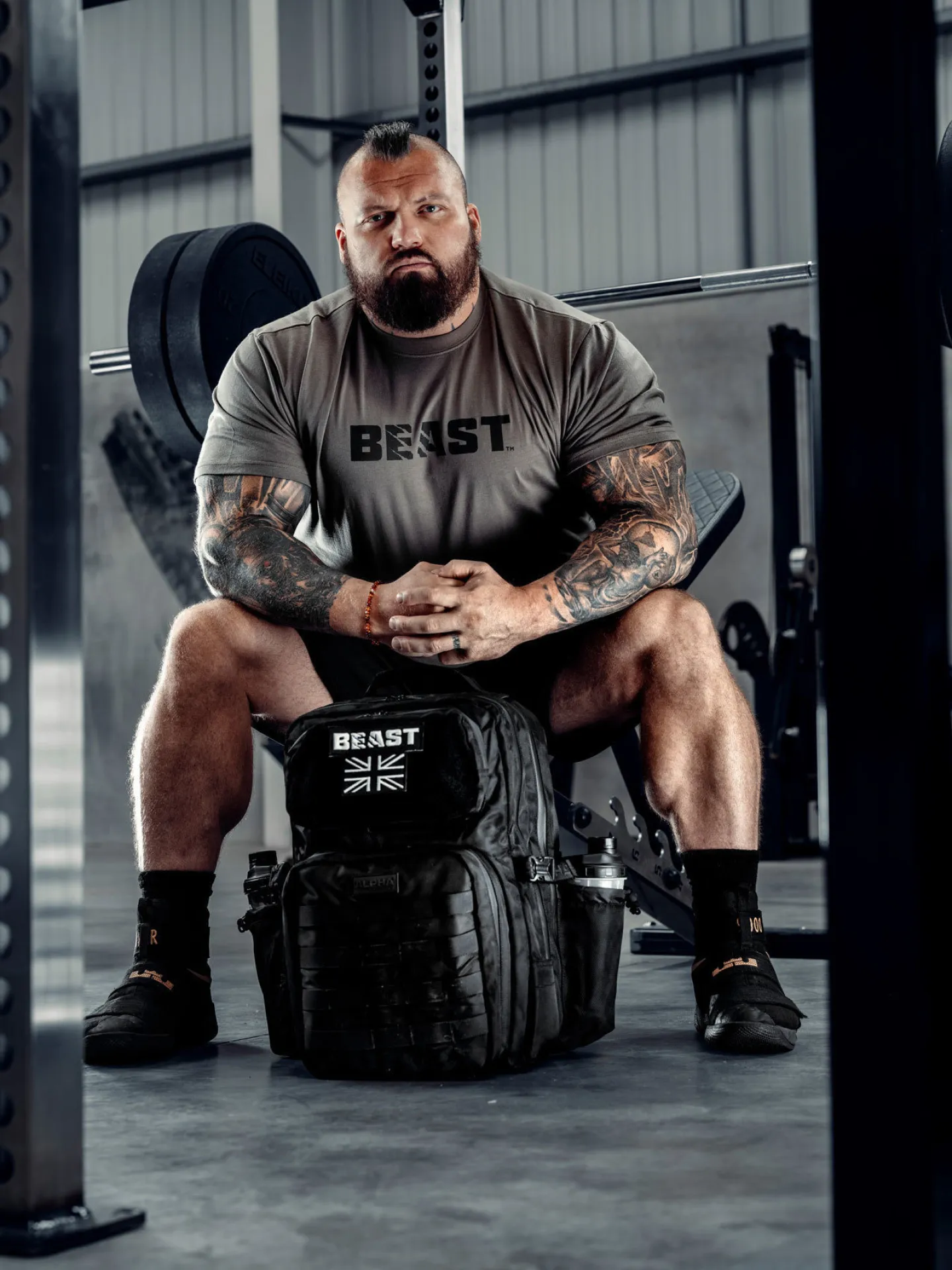 Strongman Eddie Hall seated on a gym bench with a Beast branded backpack at his feet, a loaded barbell visible behind him
