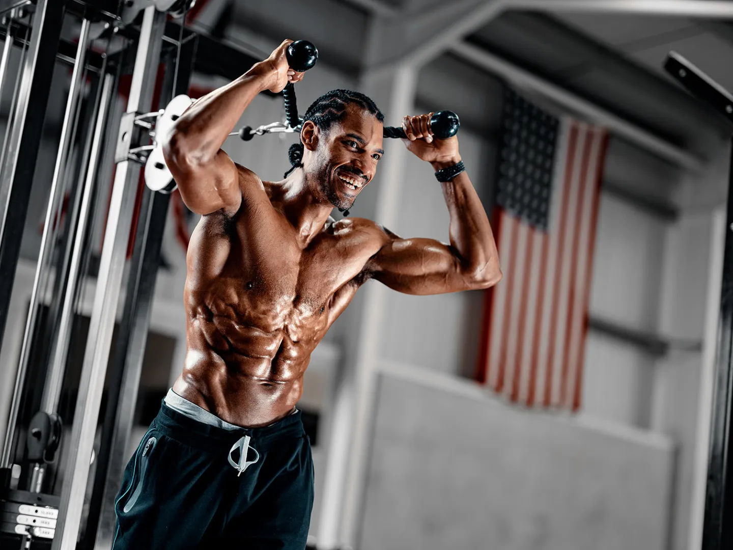 A heavily muscular man performs a cable fly exercise on a cable machine in a gym, grinning at the camera, with an American flag visible in the background