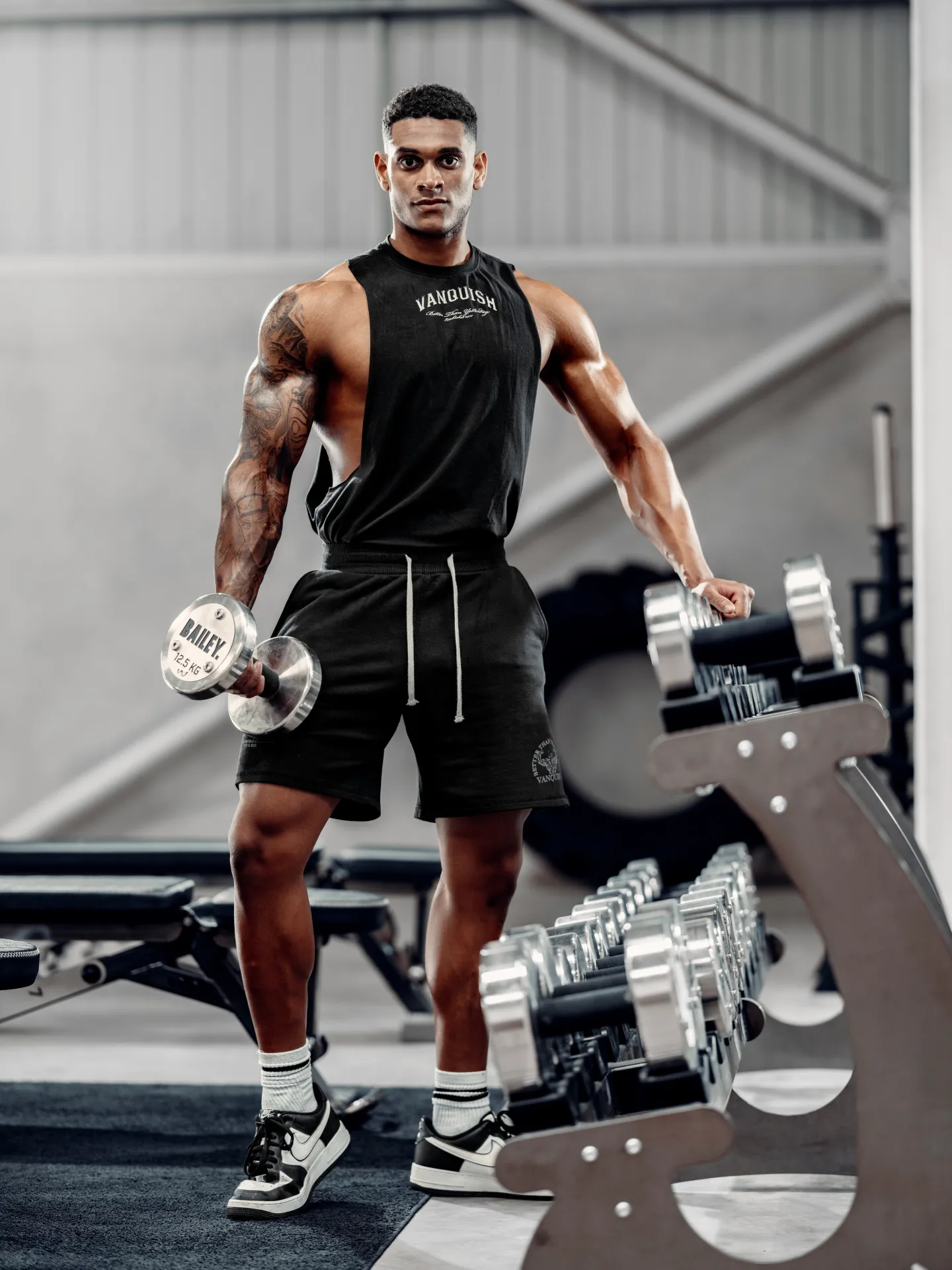 A muscular man wearing Vanquish gym wear holds a dumbbell beside a dumbbell rack in a gym studio