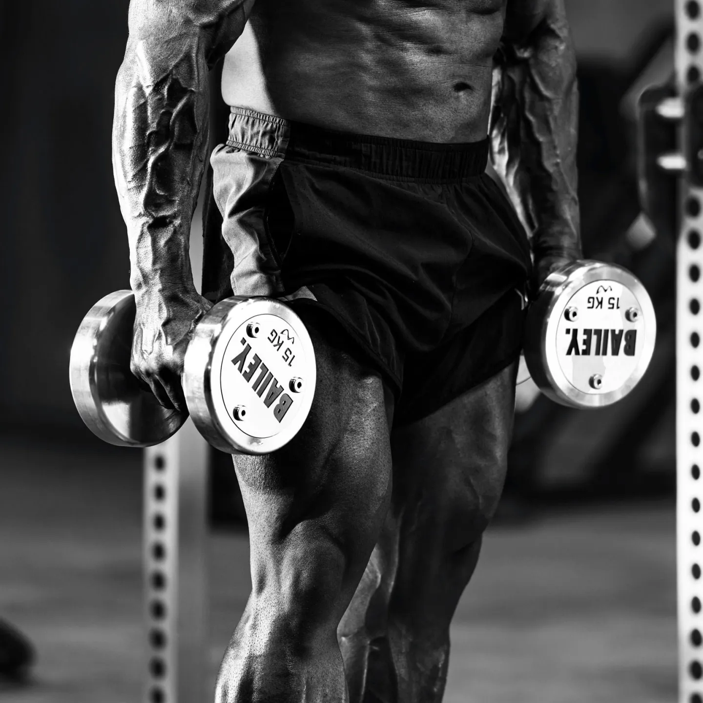 Close-up black and white photograph of a muscular mans torso and legs holding a 15kg Bailey-branded dumbbell in each hand in a gym