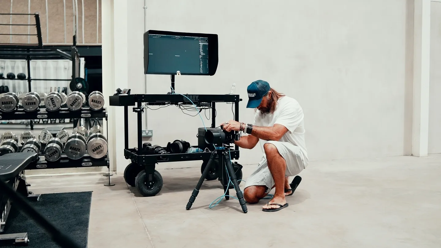 Christopher Bailey crouching to adjust a camera on a tripod inside a gym, with a monitor and equipment cart beside him and a rack of dumbbells in the background