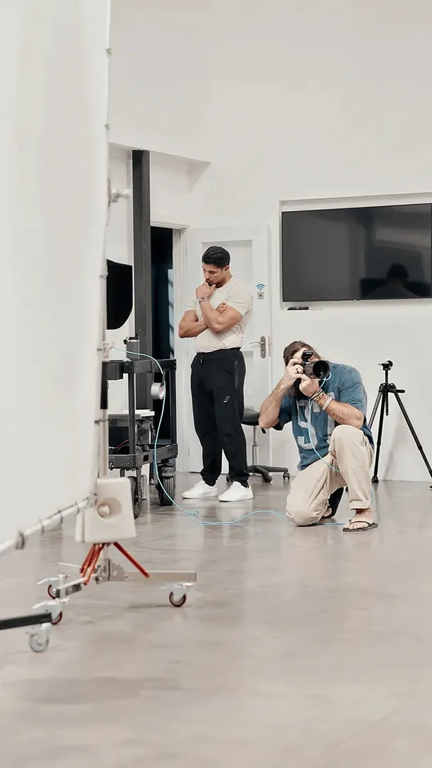 A photographer kneels on a studio floor shooting upward toward a muscular male subject standing near a white backdrop, with studio lighting equipment visible in the foreground