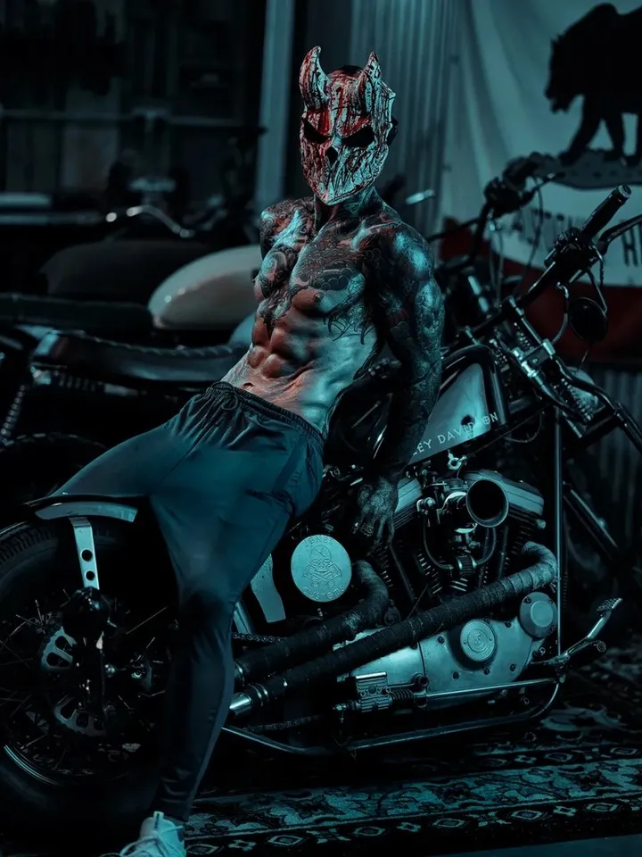 A heavily tattooed shirtless male model wearing a horned demon mask sits on a Harley-Davidson motorcycle in a dark garage setting