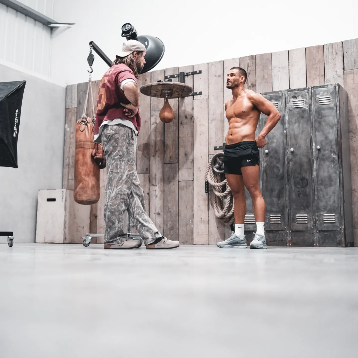 A photographer directing a lean male fitness model in a studio set with gym lockers, a heavy bag and studio lighting equipment visible