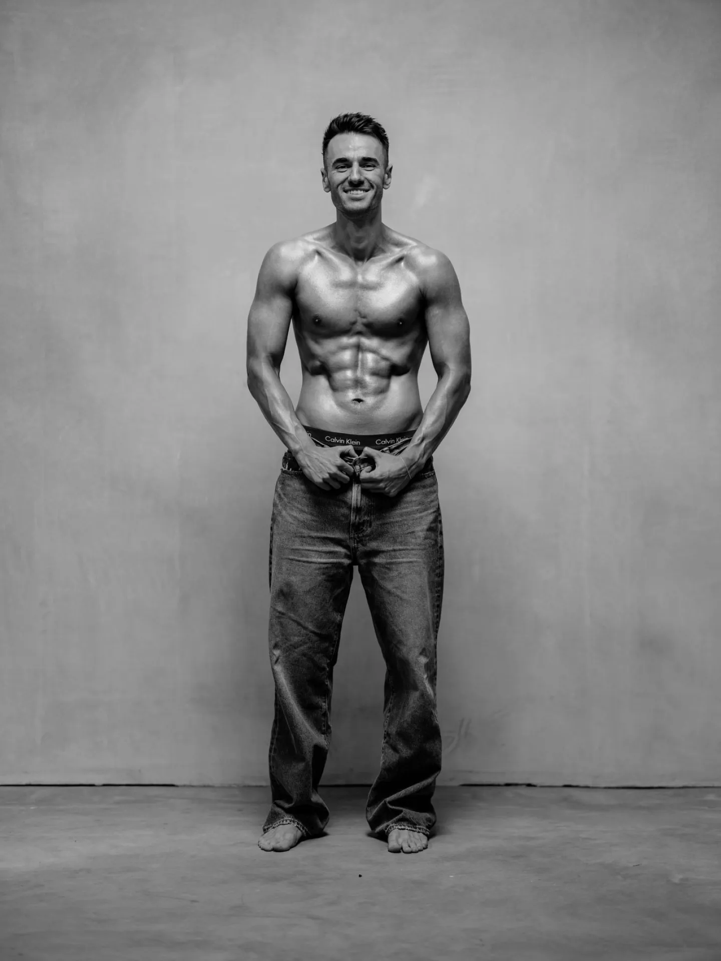 Black and white full length studio portrait of a muscular man shirtless in jeans standing and smiling at the camera