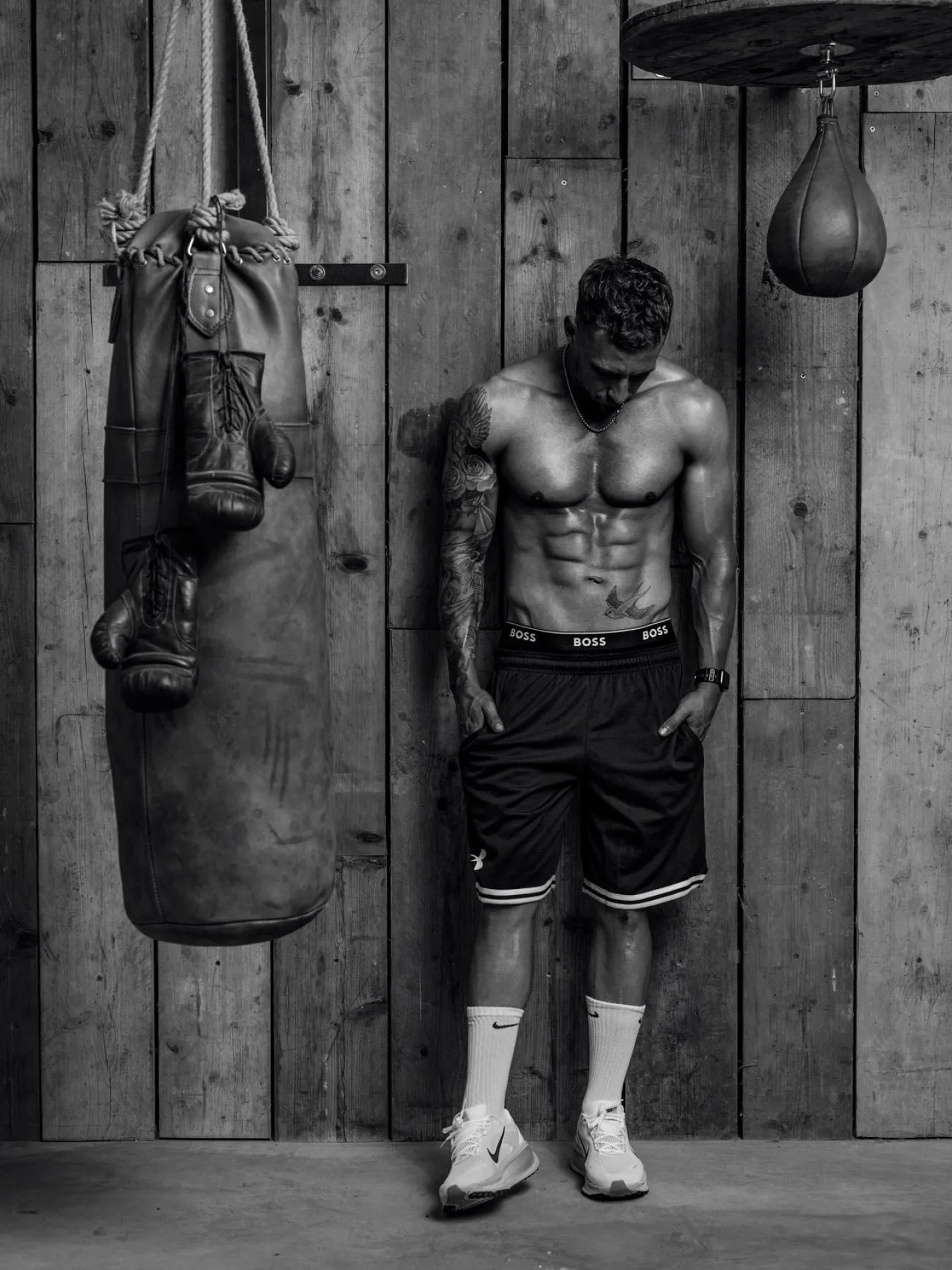 A muscular tattooed man stands shirtless against a wooden wall in a boxing gym, a heavy bag and speed ball hanging nearby, shot in black and white