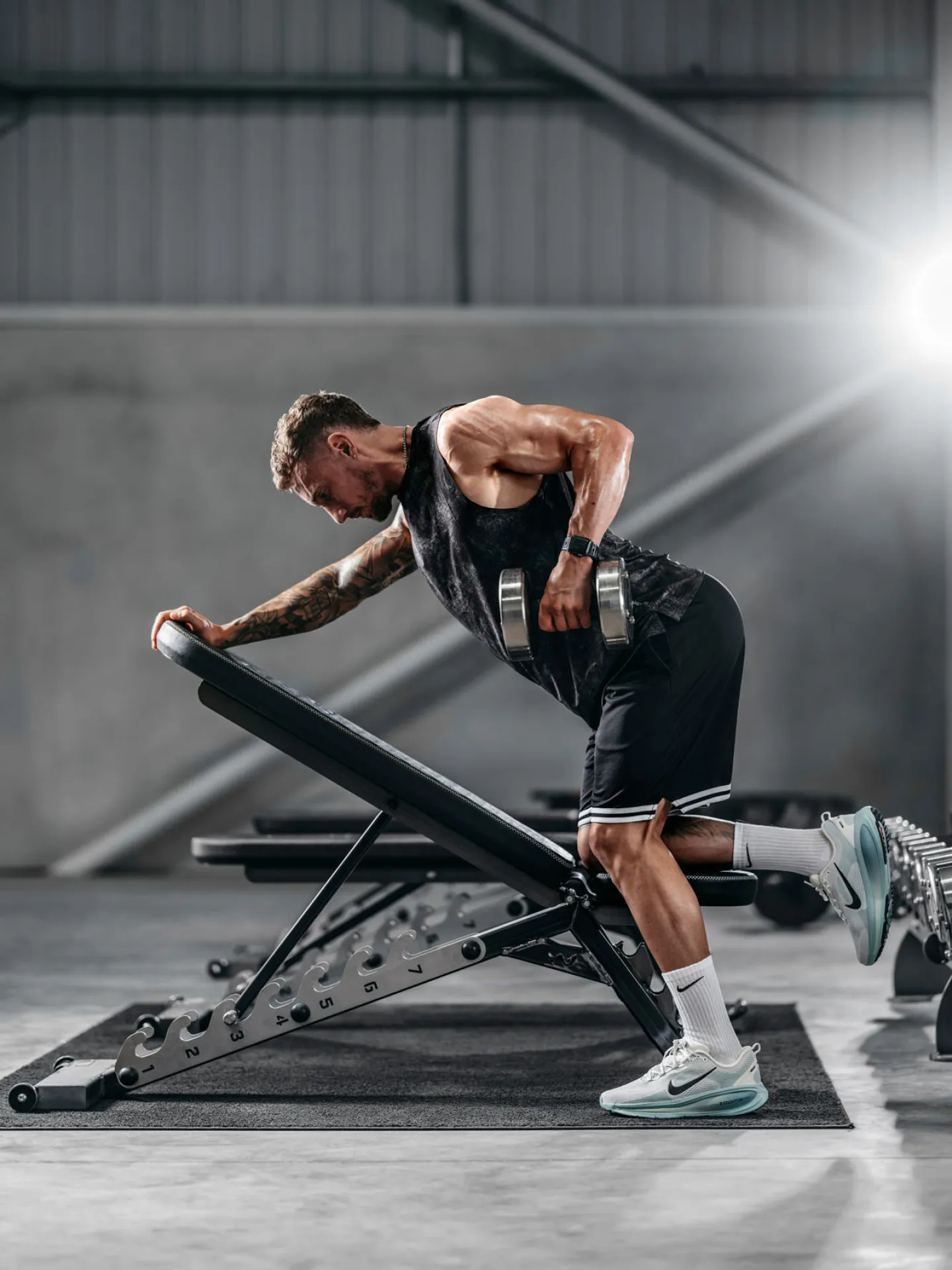 A tattooed male athlete performs a dumbbell row on an incline bench in a large industrial gym, leaning forward with a dumbbell raised to his side