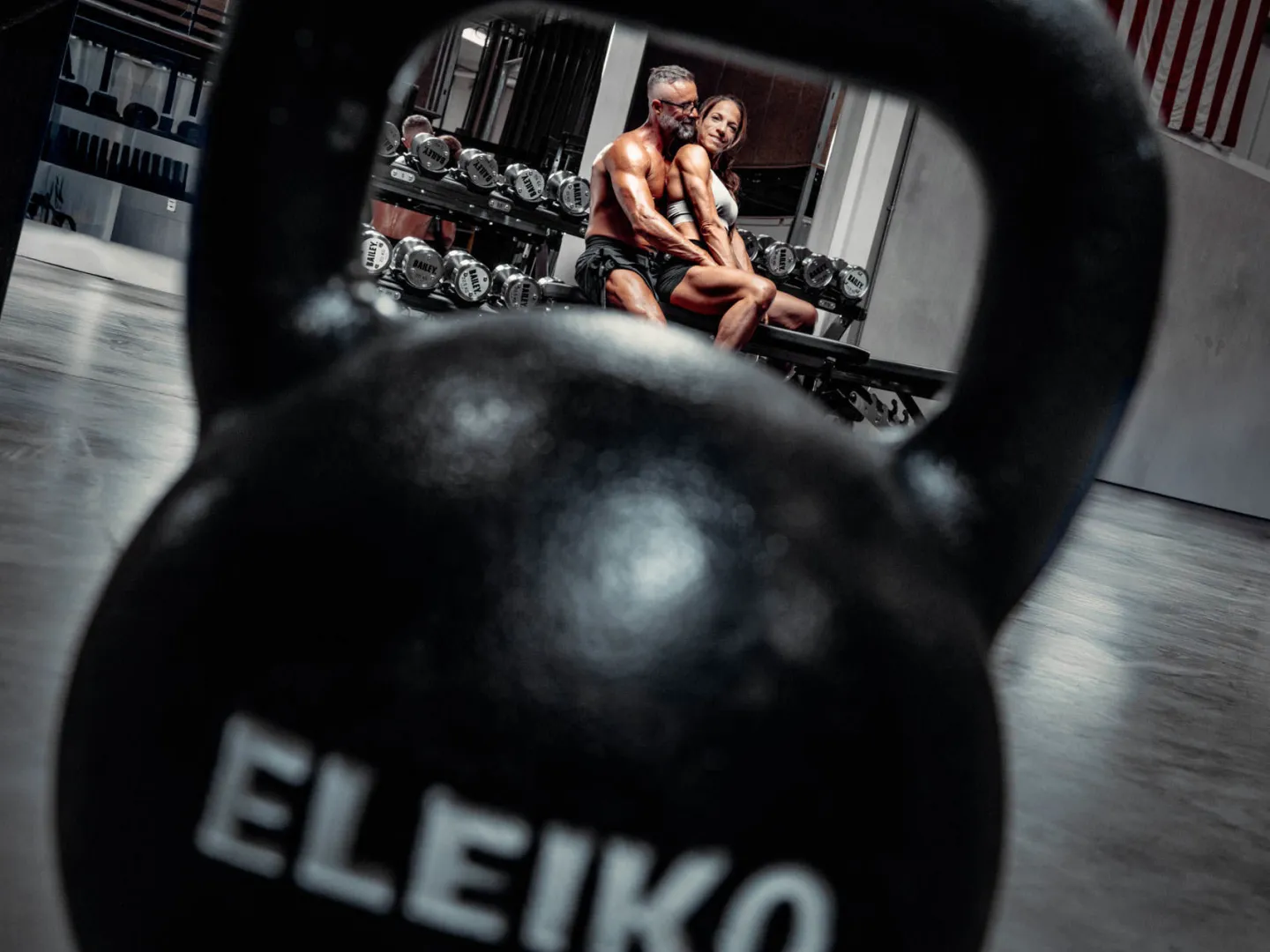A muscular man and a lean woman sit close together on a gym bench framed through the handle of a large Eleiko kettlebell in the foreground, with a dumbbell rack visible behind them