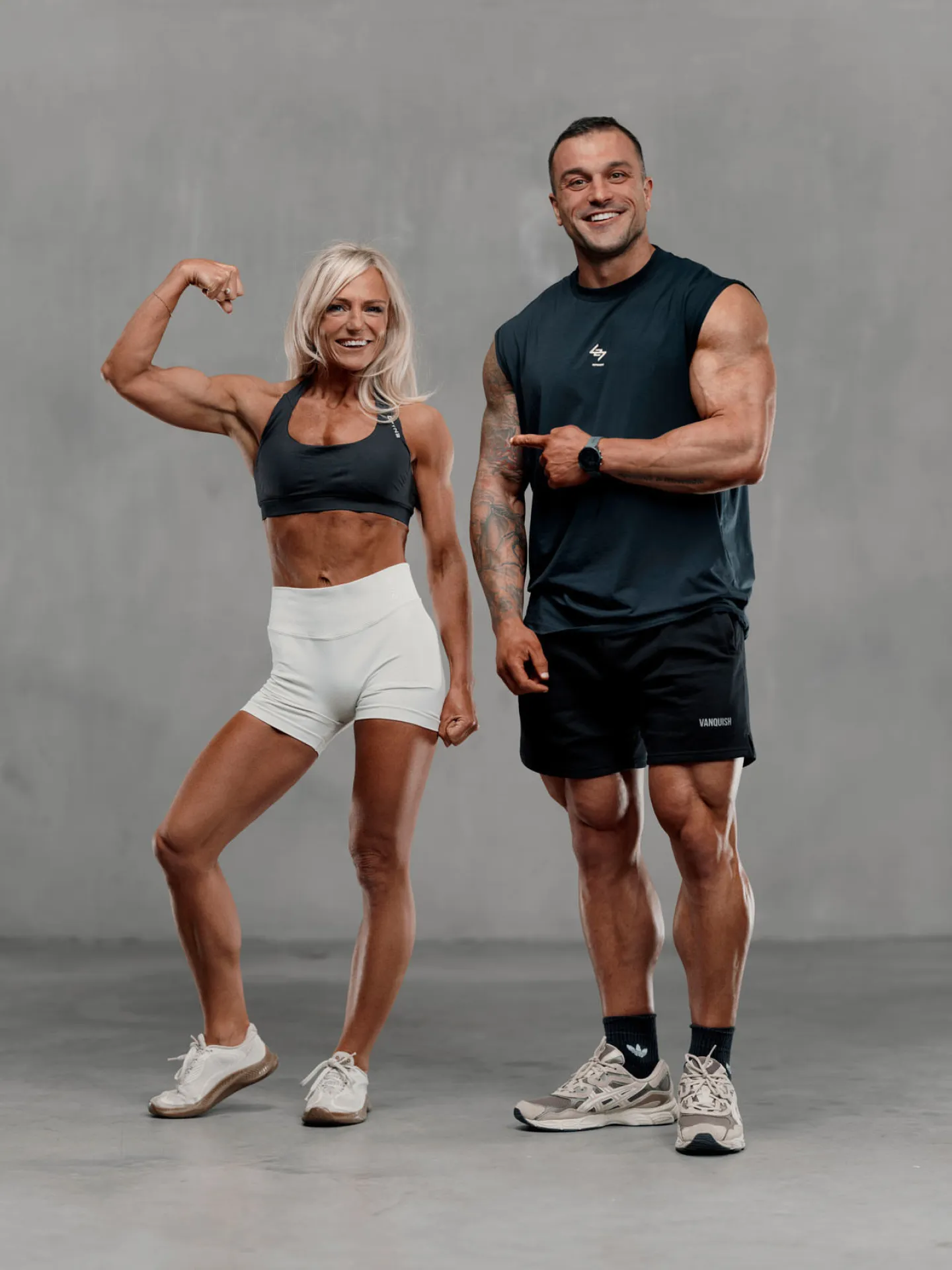 A muscular woman flexing her bicep and a tall muscular man standing beside her, both smiling, against a grey studio backdrop