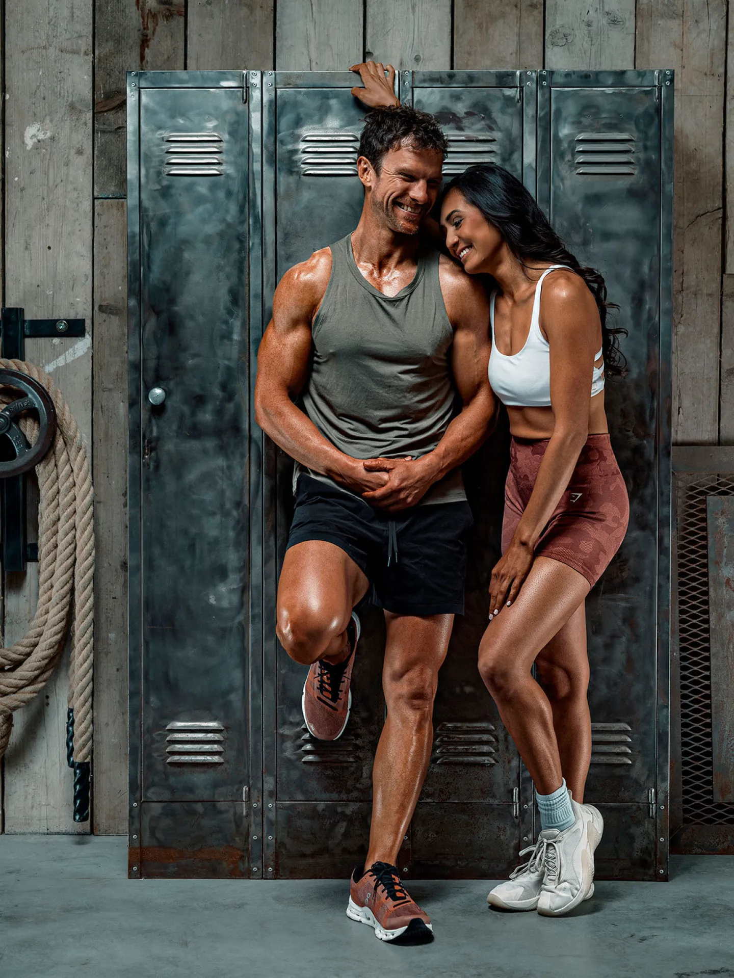 A man and woman in gym wear lean together smiling in front of metal lockers in an industrial gym setting