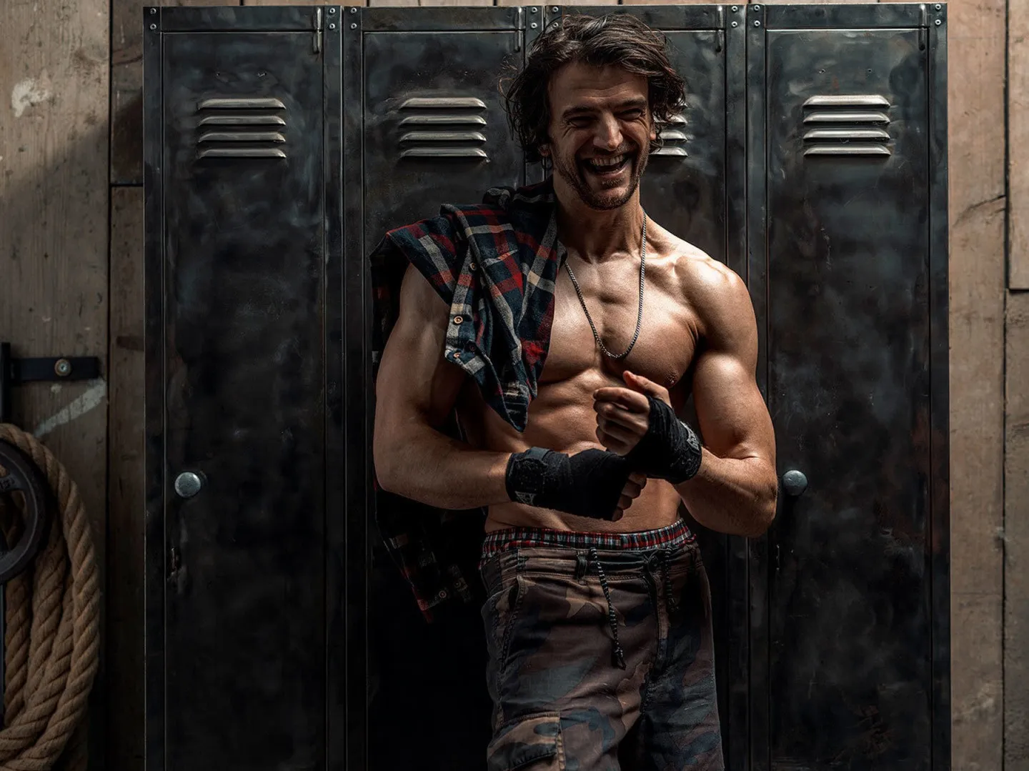 A muscular man with dark curly hair laughs while leaning against metal gym lockers, wearing an open flannel shirt and boxing hand wraps