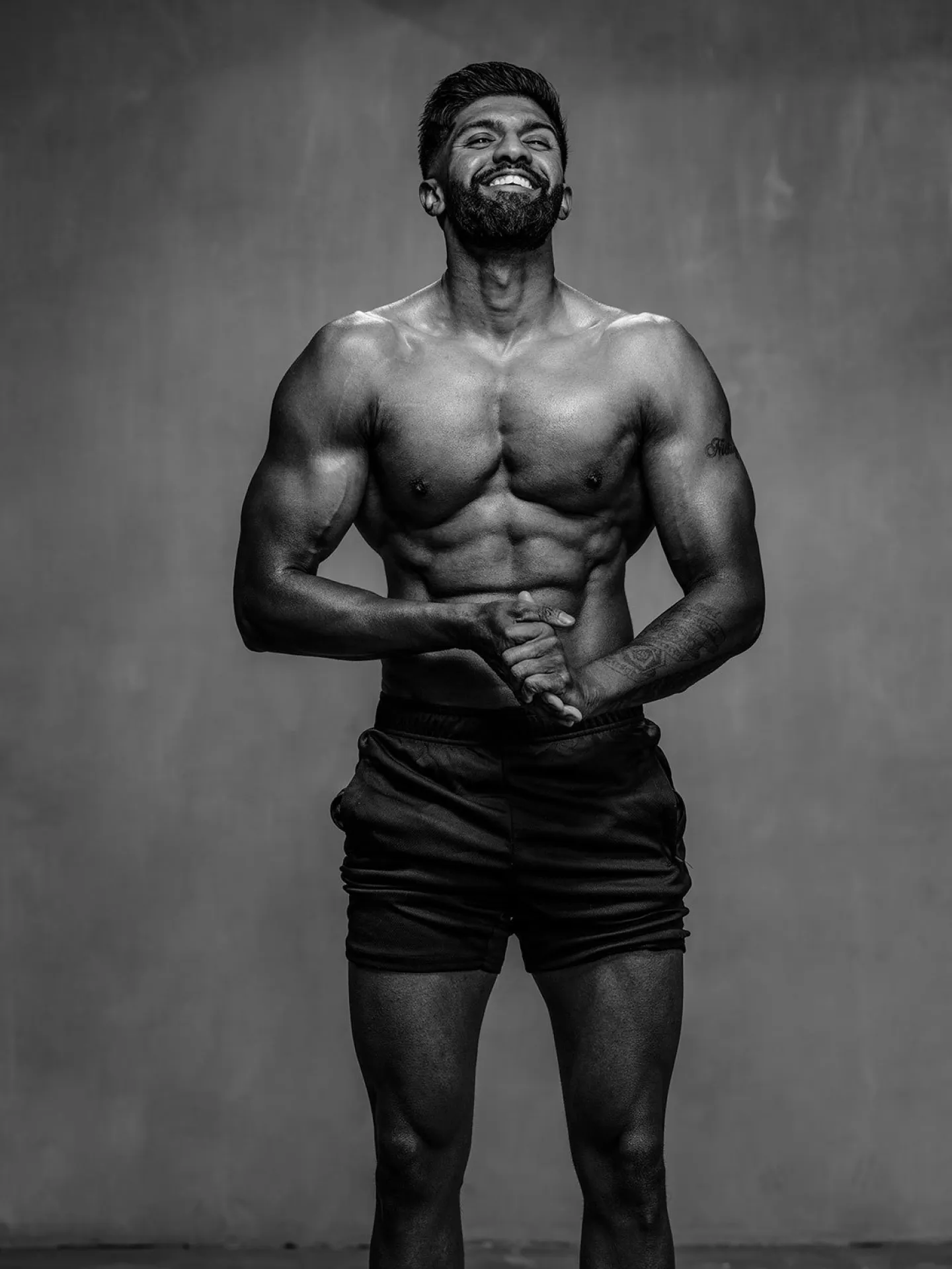 Black and white studio portrait of a muscular bearded man smiling with his hands clasped at his waist