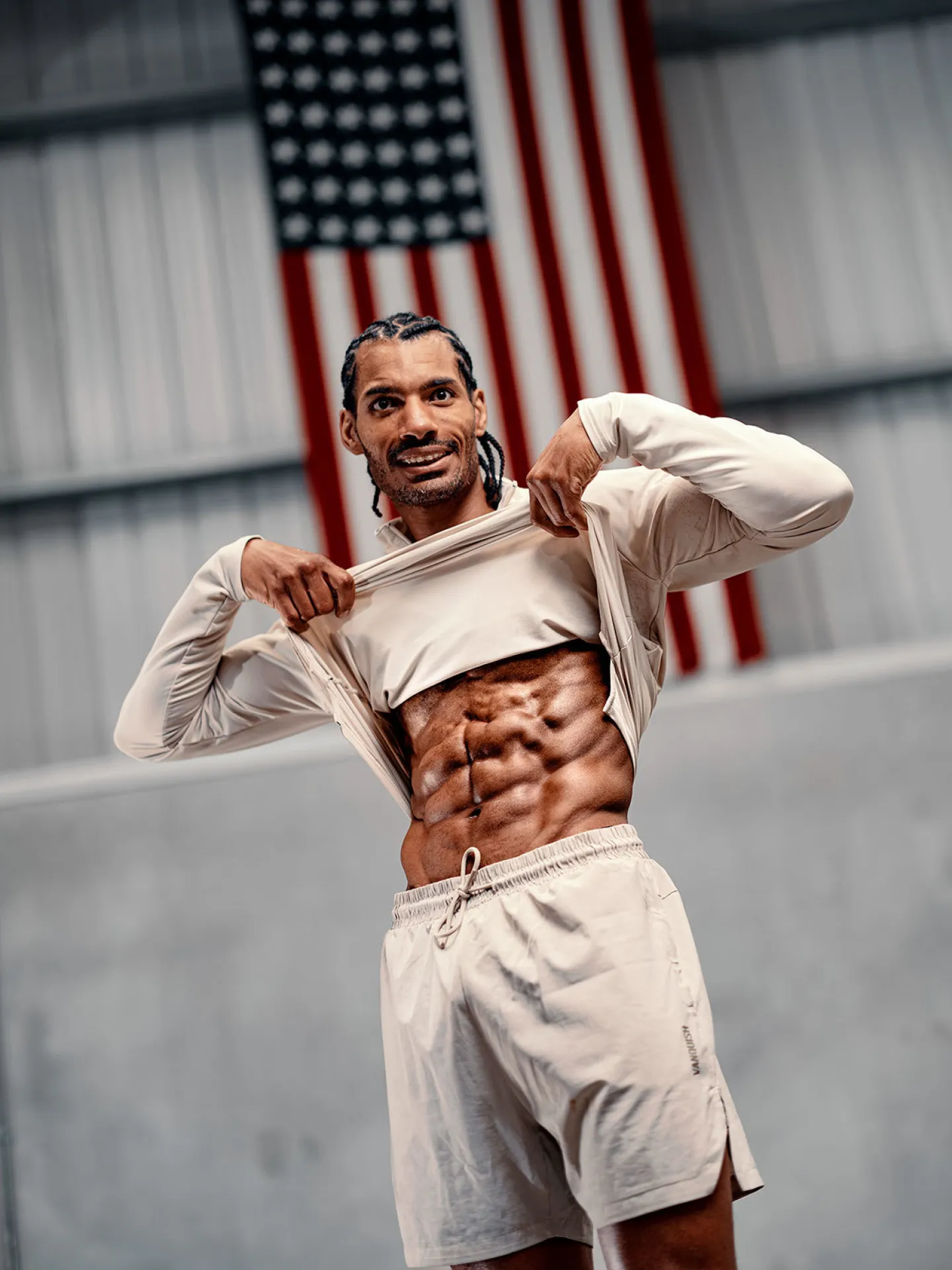A lean muscular man lifts his long-sleeve top to reveal his abdomen while smiling, with a large American flag hanging on a metal wall behind him