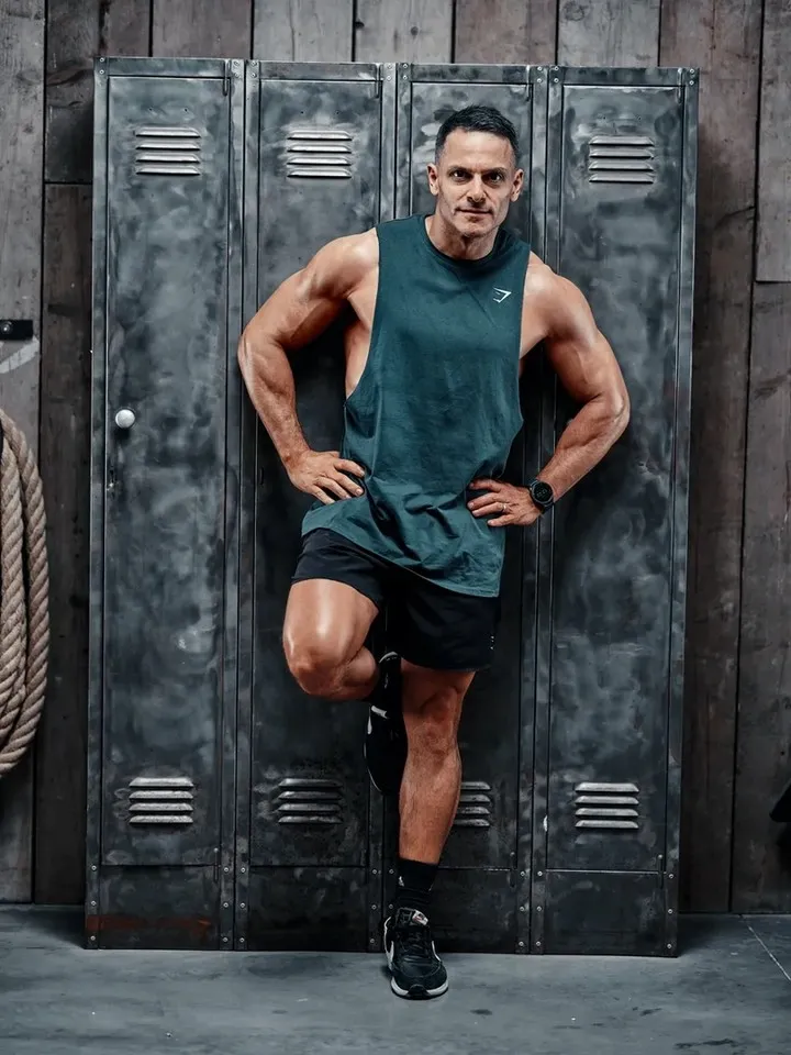 A muscular man in a teal Gymshark vest and black shorts stands leaning against metal gym lockers with one foot raised, looking directly at the camera