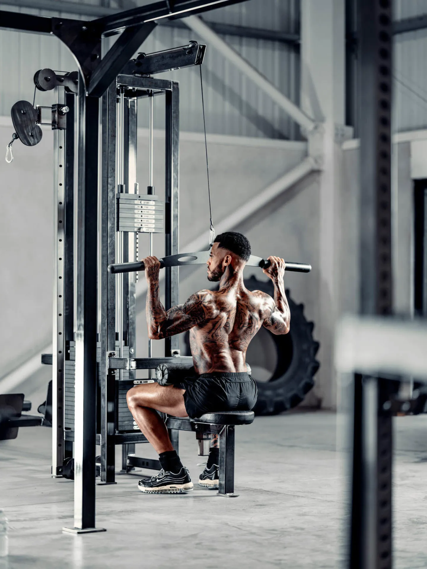 A heavily tattooed muscular man performs a lat pulldown on a cable machine in a large industrial gym, photographed from behind and to the side