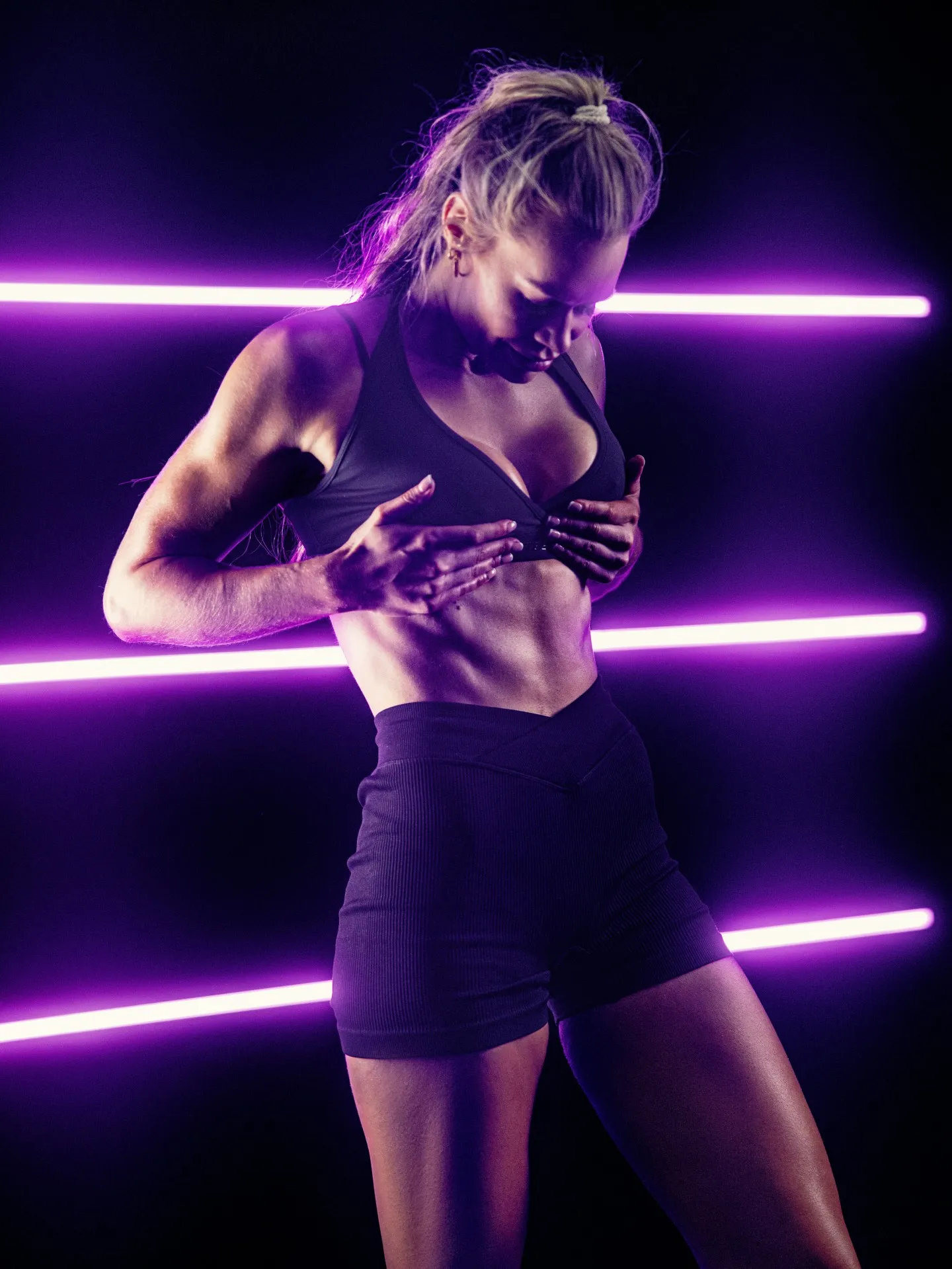 A athletic blonde woman in dark sports bra and shorts poses against a black background lit by horizontal purple neon tubes