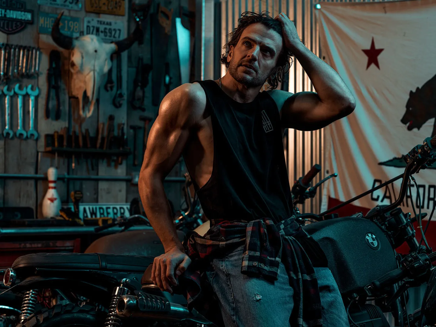 A muscular man in a black sleeveless top and jeans leans against a vintage BMW motorcycle in a dimly lit garage decorated with American licence plates, tools, a bull skull and a California Republic flag