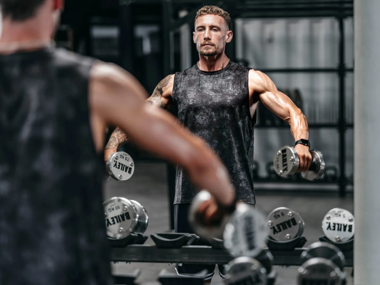 A muscular man in a sleeveless top holds dumbbells branded Bailey in a gym, photographed from behind a rack of weights with a blurred foreground