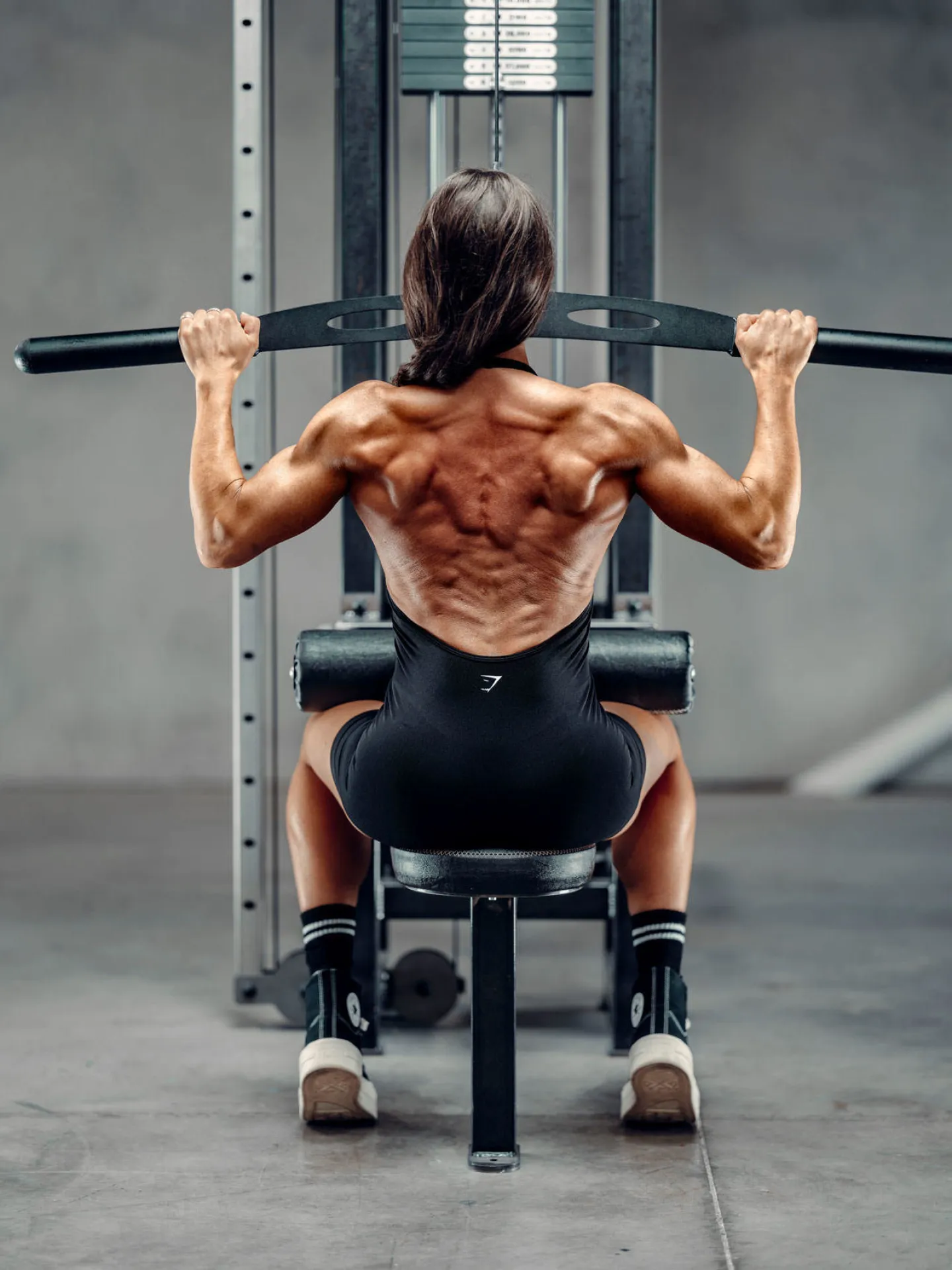 A muscular woman seen from behind performing a lat pulldown on a cable machine in a gym, wearing black Gymshark sportswear