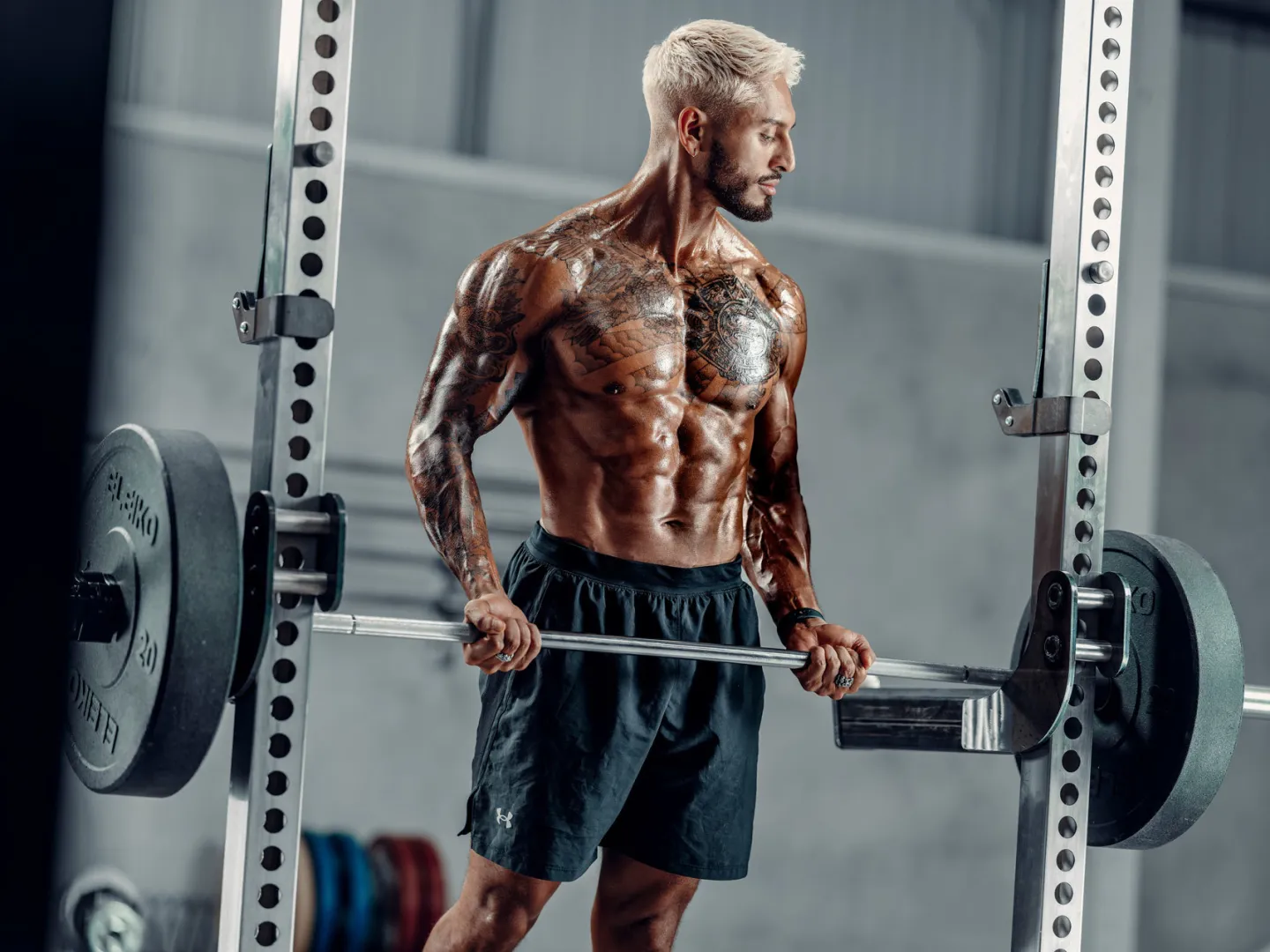 A heavily tattooed muscular man with bleached hair holds a loaded barbell inside a squat rack in a gym