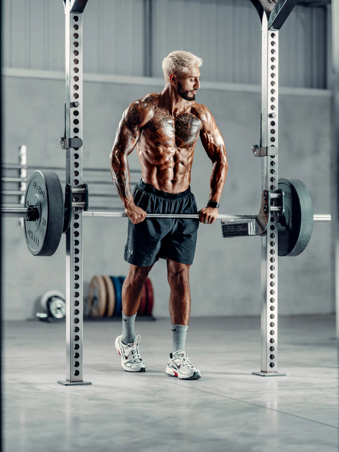 A lean muscular tattooed man with bleached hair holds a loaded barbell inside a power rack in a large gym