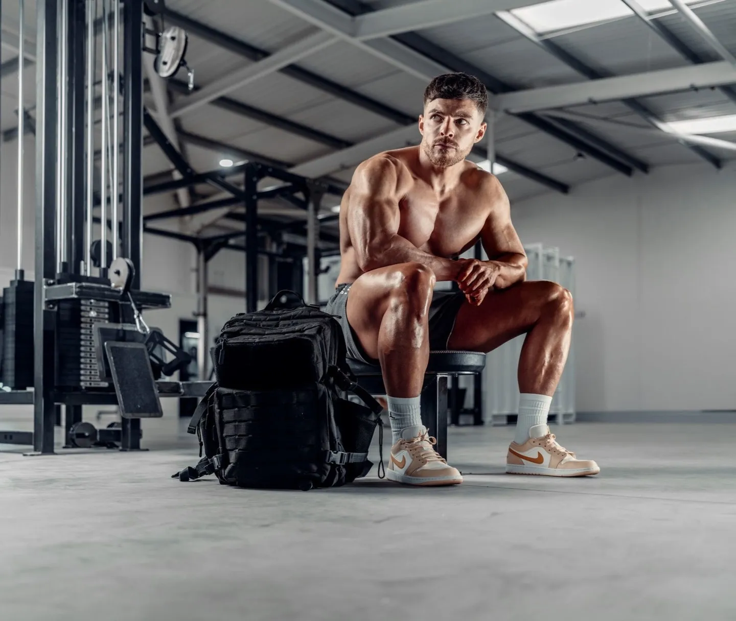 A muscular man sits on a gym bench in a large training facility, shirtless, leaning forward with hands clasped, a black tactical backpack on the floor in front of him