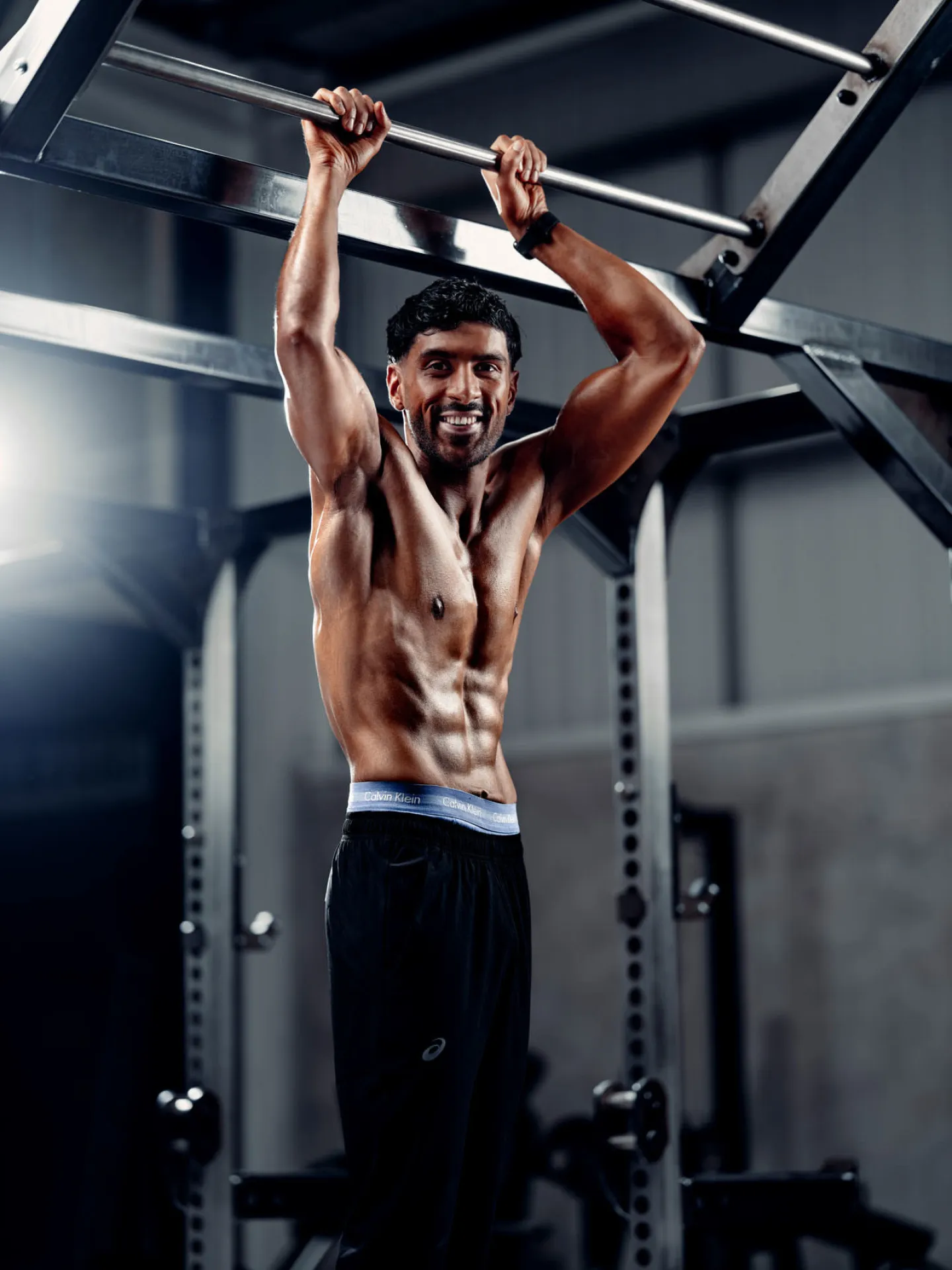 A lean muscular man hangs smiling from a pull-up bar in a gym, shirtless, wearing black shorts with a Calvin Klein waistband, with a power rack visible behind him