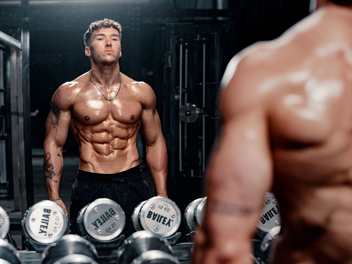 A muscular shirtless man with tattoos stands at a dumbbell rack in a gym, reflected in a mirror behind him. The dumbbells are branded Bailey.