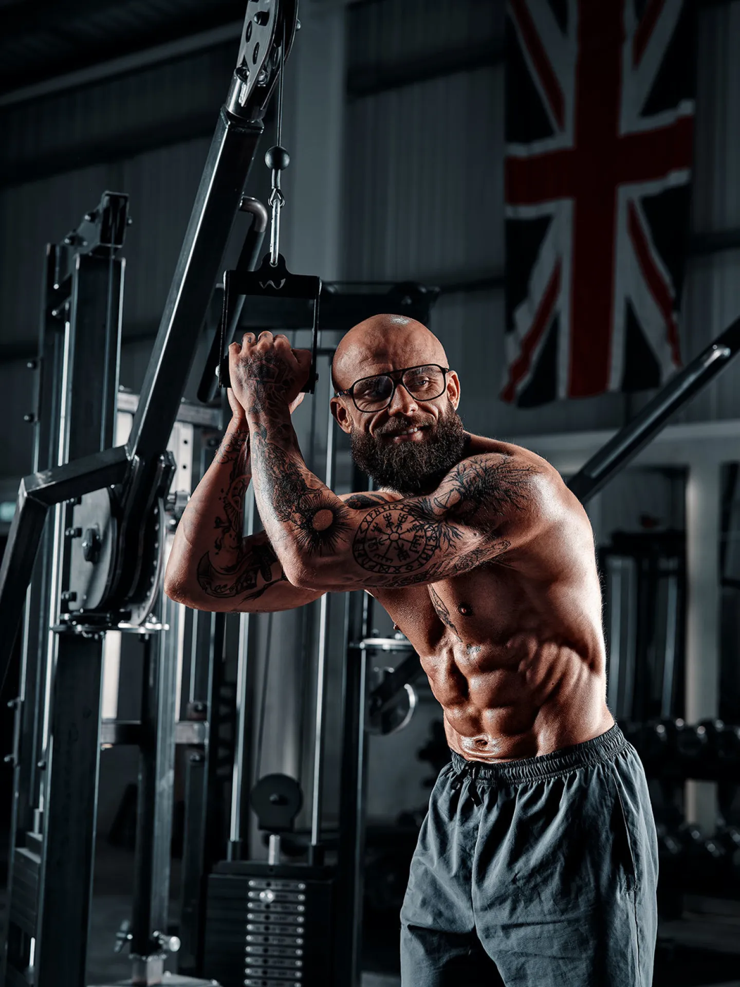 A heavily tattooed bald man with a beard and glasses trains on a cable machine in a gym with a Union Jack flag on the wall behind him