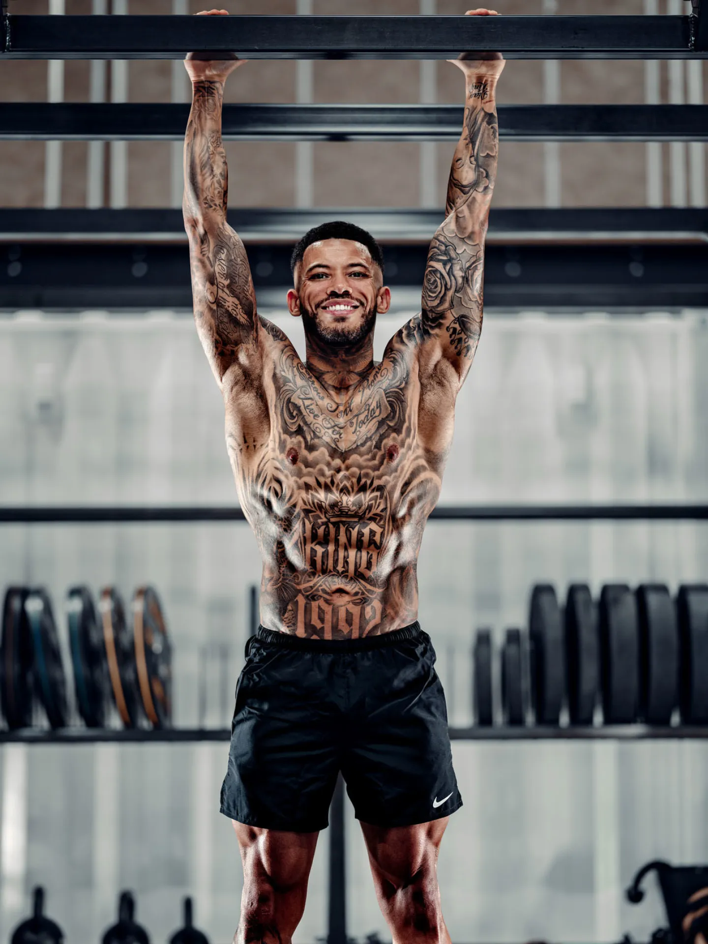 A heavily tattooed man hangs from a pull-up bar in a gym, smiling at the camera, with weight plates visible on racks behind him