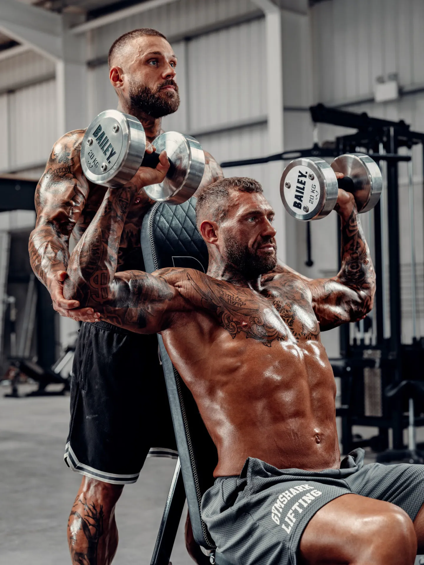 Two heavily tattooed muscular men holding 20kg Bailey dumbbells in a gym, one seated on an incline bench performing a shoulder press, the other standing behind him curling the weights