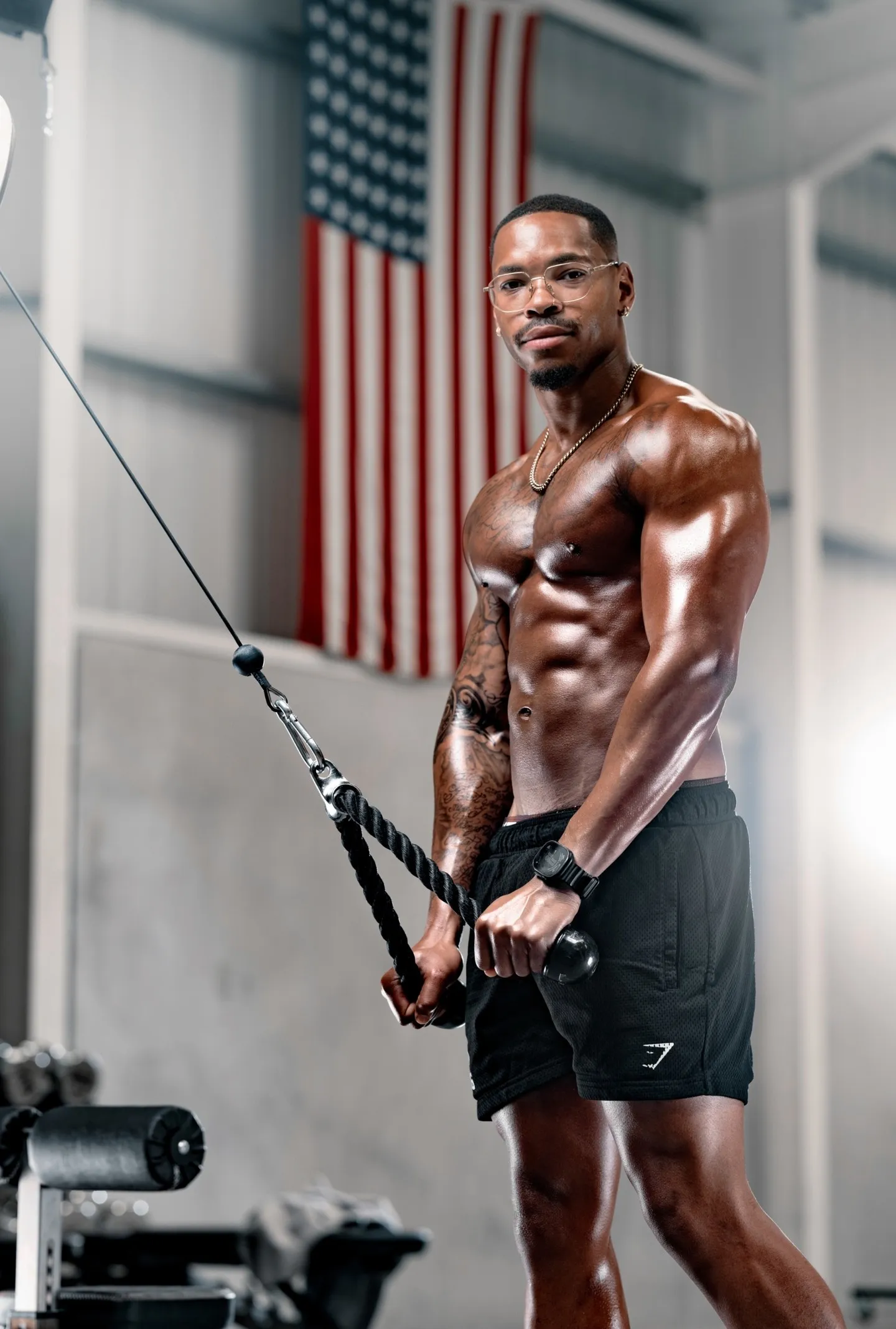 A muscular man holds a cable machine rope attachment in a gym with an American flag on the wall behind him