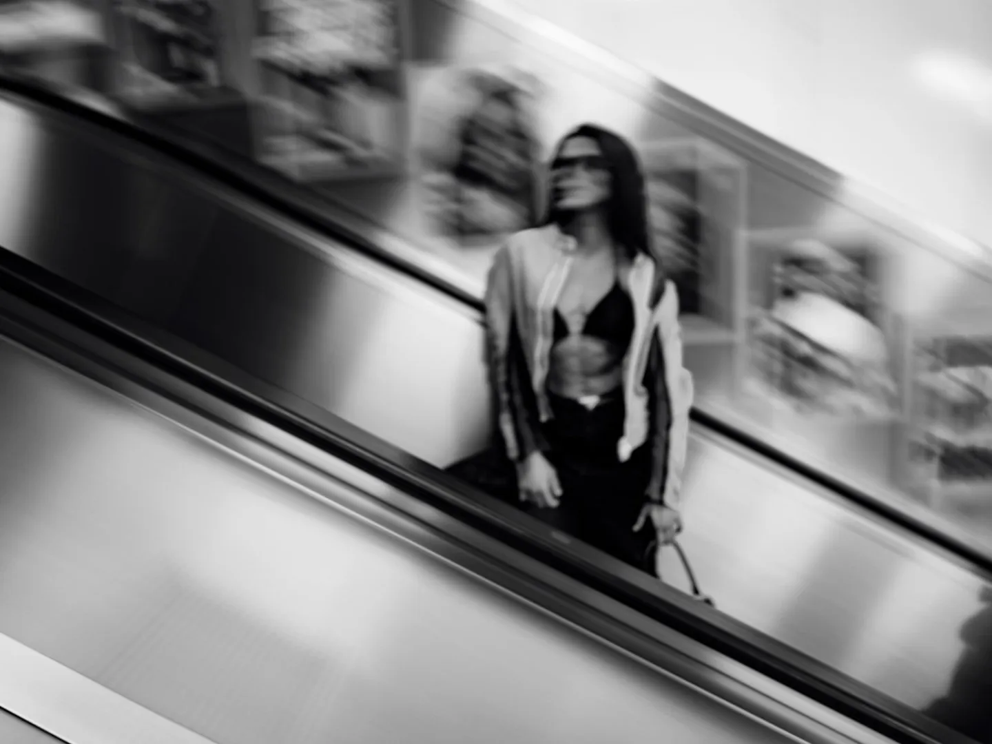 Black and white motion-blurred photograph of a woman riding an escalator in a shopping centre, wearing sunglasses and carrying a handbag
