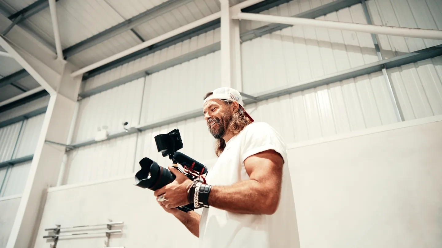 Fitness photographer Christopher Bailey smiling while holding a camera on a gimbal stabiliser inside a large white industrial studio space