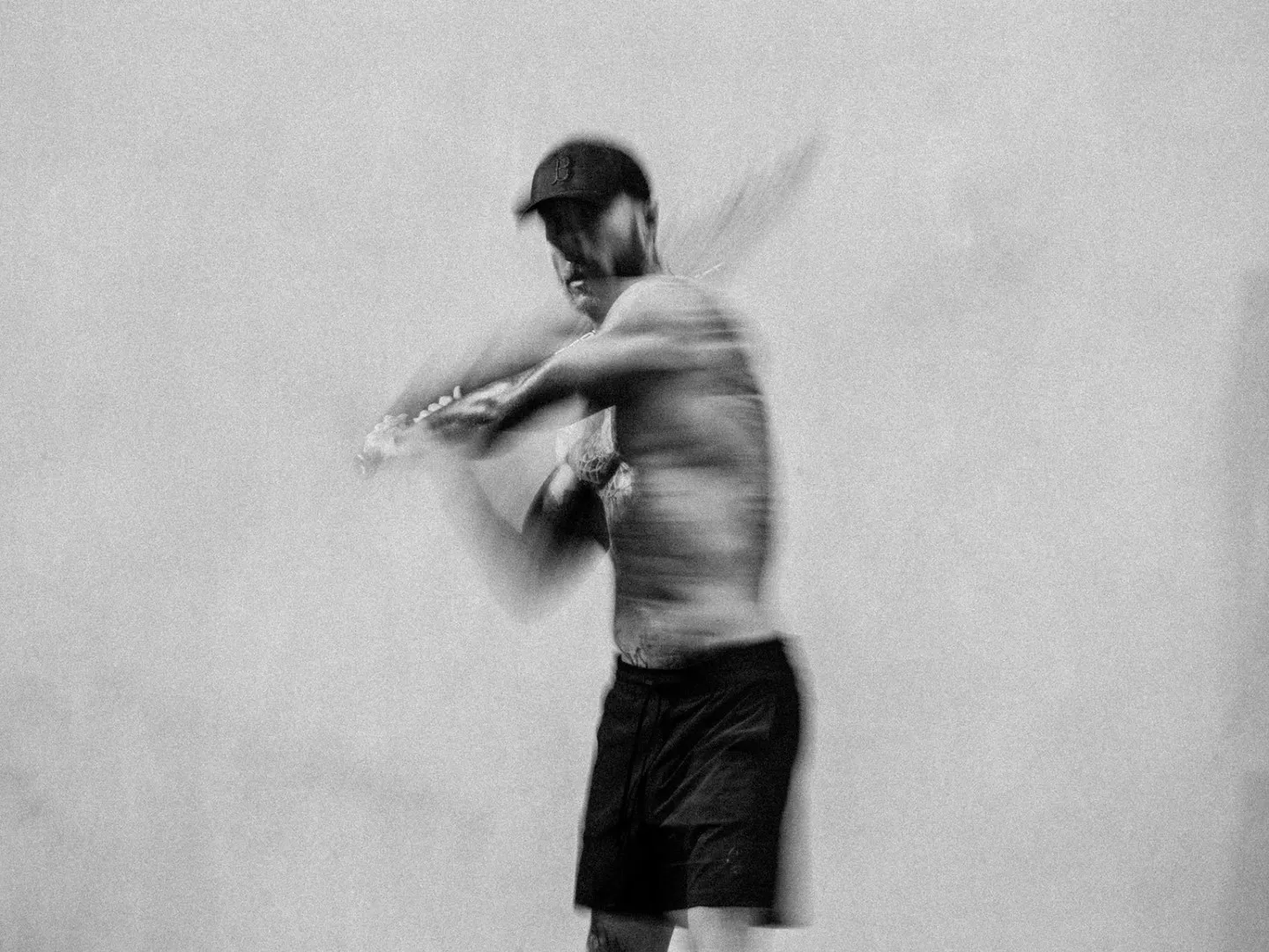 Black and white motion-blurred photograph of a man mid-swing with a baseball bat, wearing a cap and shorts, against a plain light background