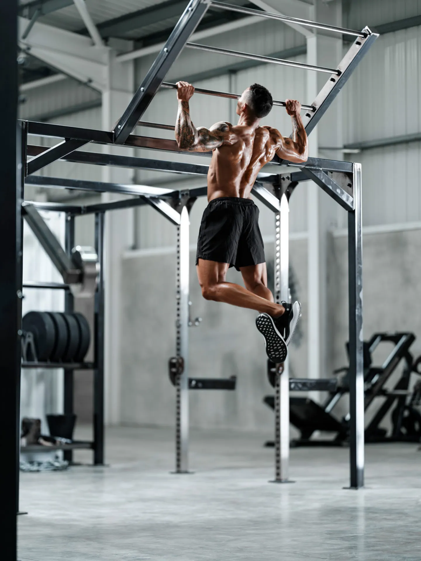 A muscular tattooed man performs a pull-up on a steel rig in a large industrial gym, photographed from behind and below