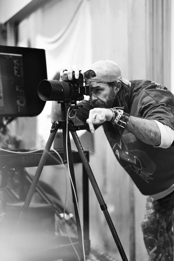 Black and white photograph of a bearded man looking through a camera on a tripod in a studio setting with a monitor visible to the left