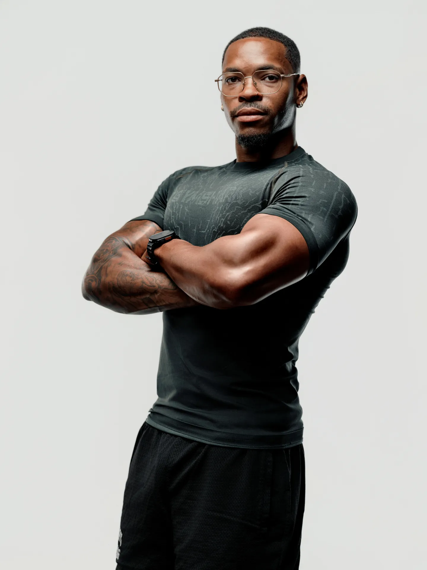 A muscular man with tattooed arms crossed, wearing wire-framed glasses and a dark fitted t-shirt, photographed against a plain light grey background