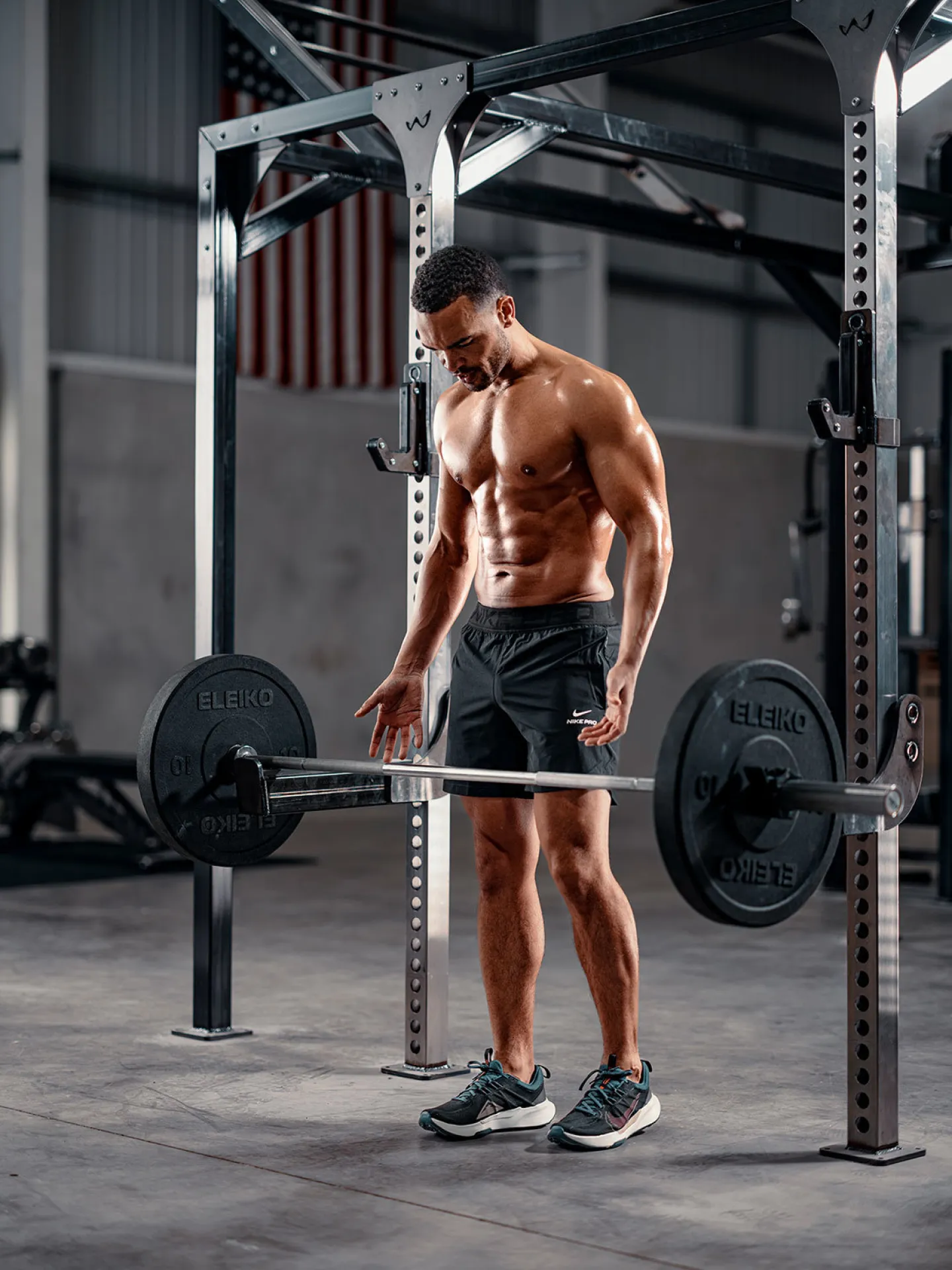 A lean muscular man stands shirtless at a loaded Eleiko barbell inside a power rack in an industrial gym