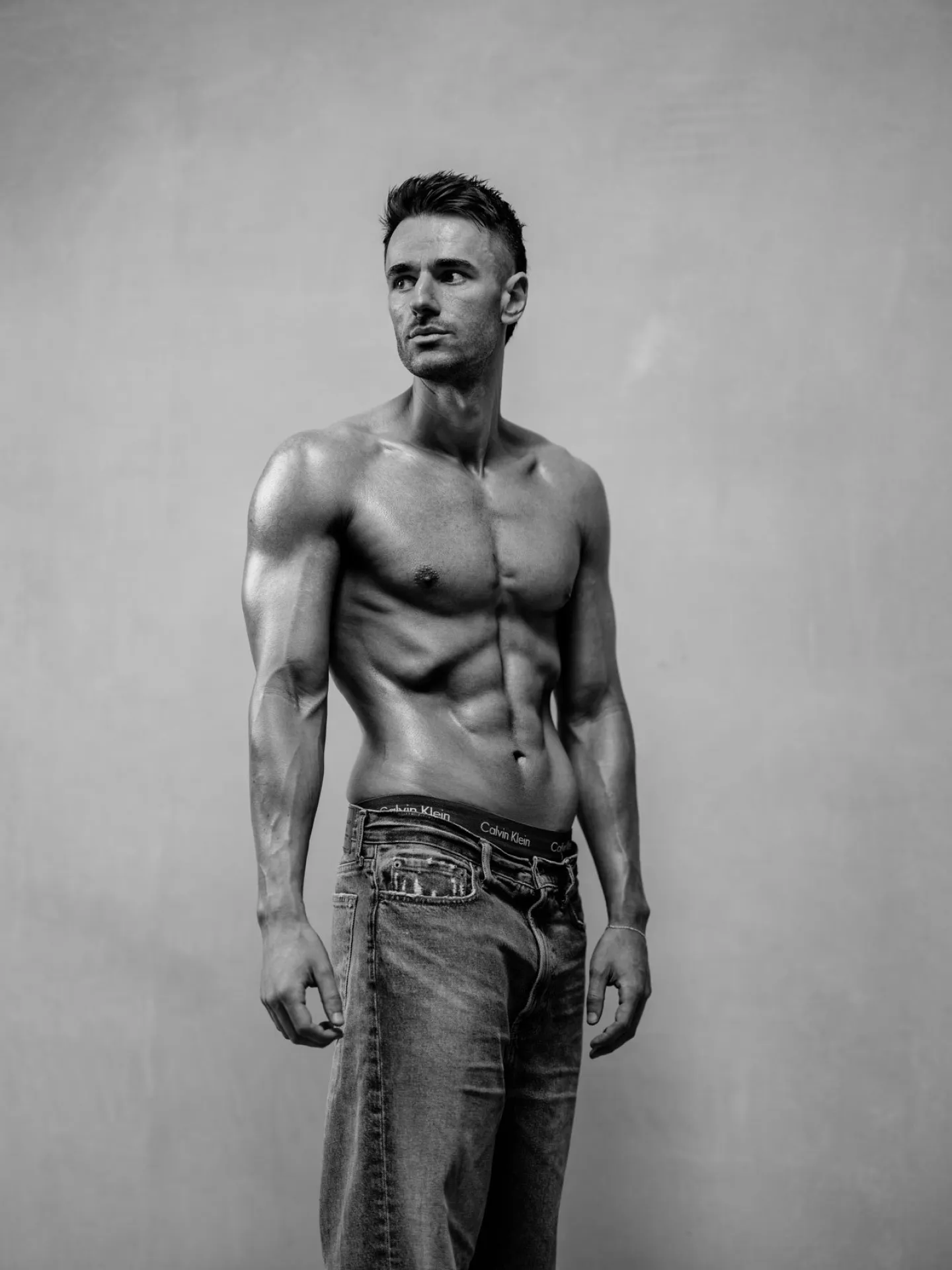 Black and white studio portrait of a lean muscular man shirtless in jeans looking to one side against a plain background