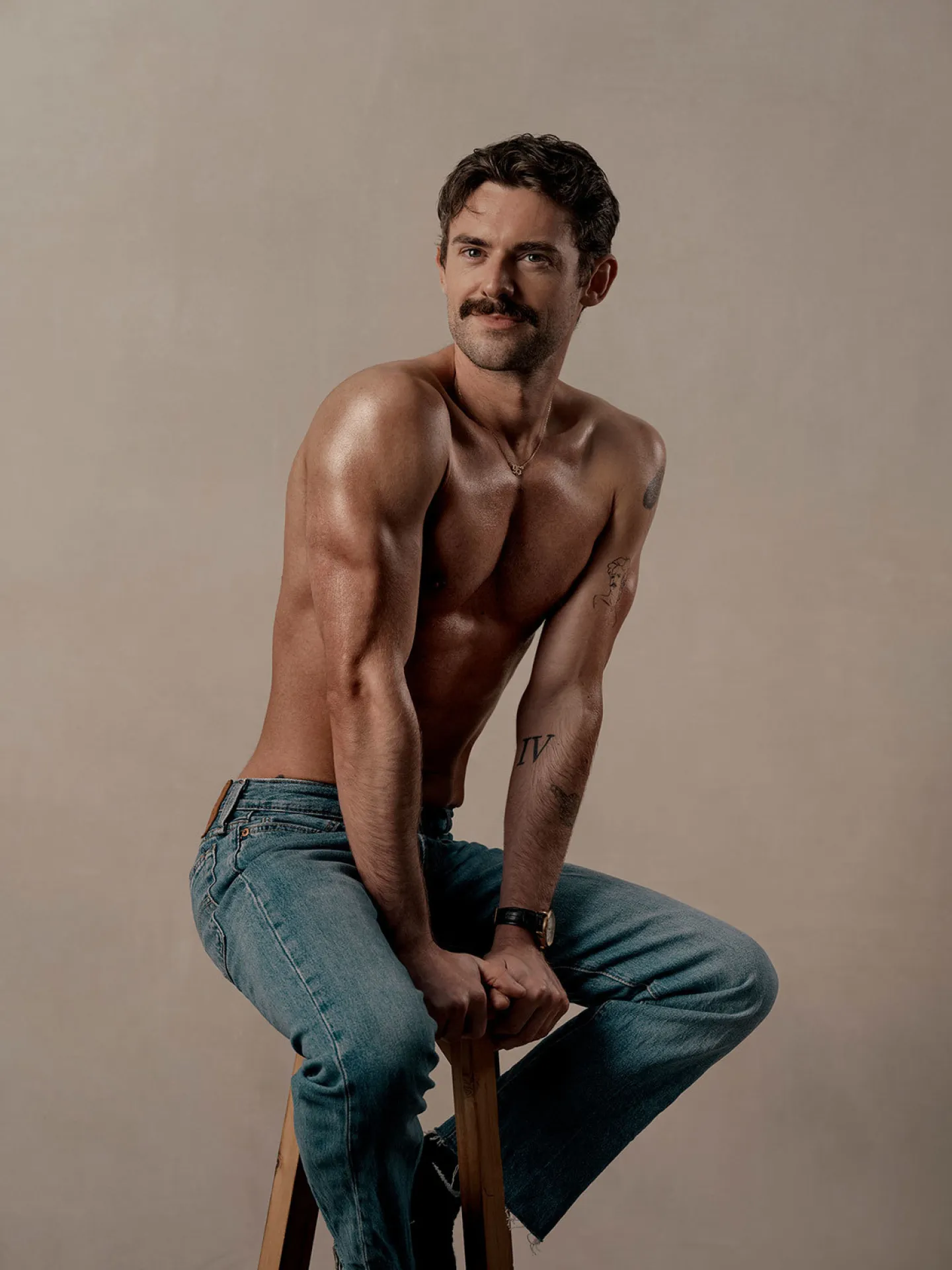 A muscular man with a moustache sits shirtless on a wooden stool in a studio, wearing jeans and a watch, looking toward the camera