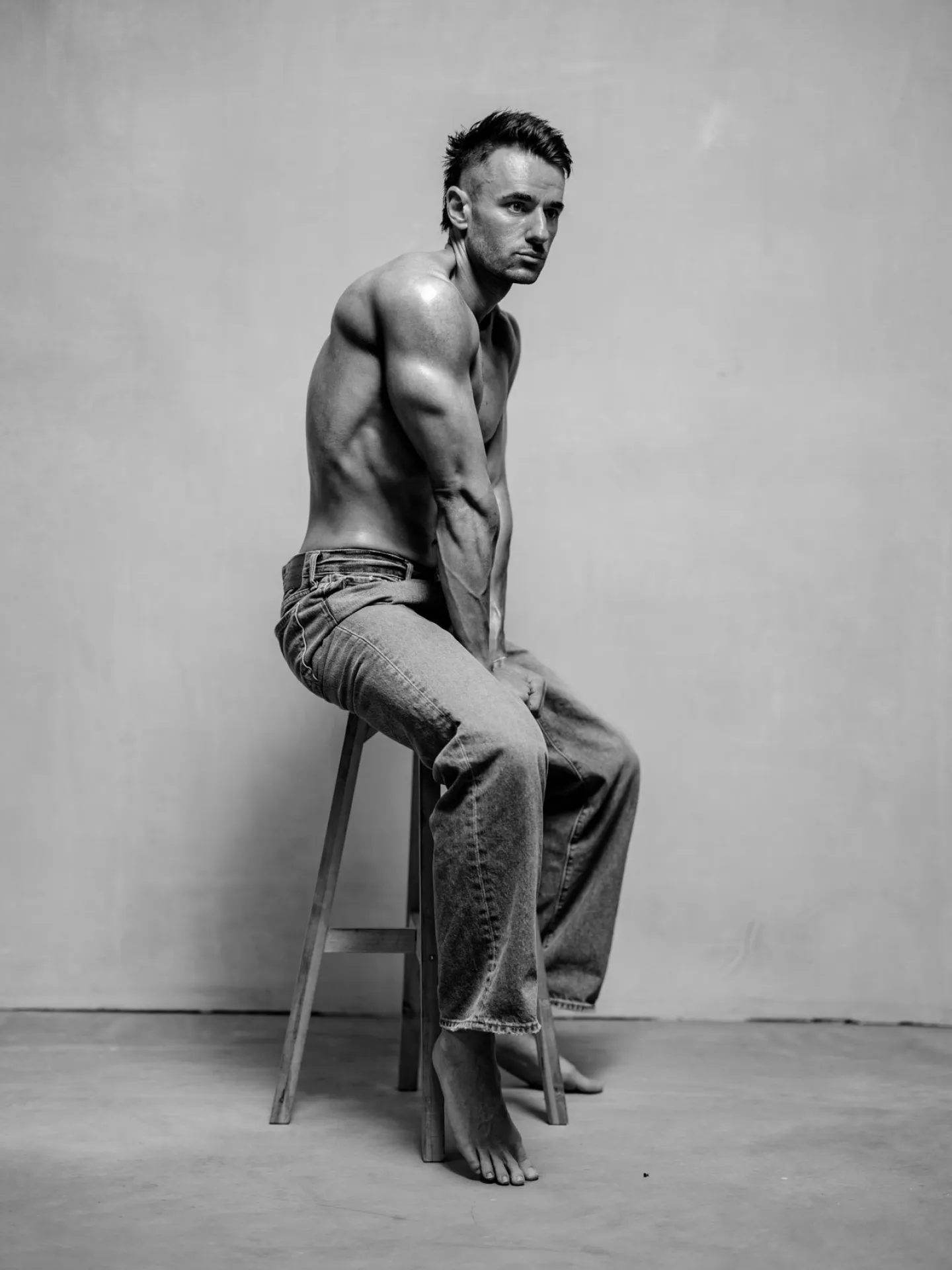 Black and white studio portrait of a muscular man seated on a wooden stool, shirtless and barefoot, wearing jeans, looking back over his shoulder toward the camera
