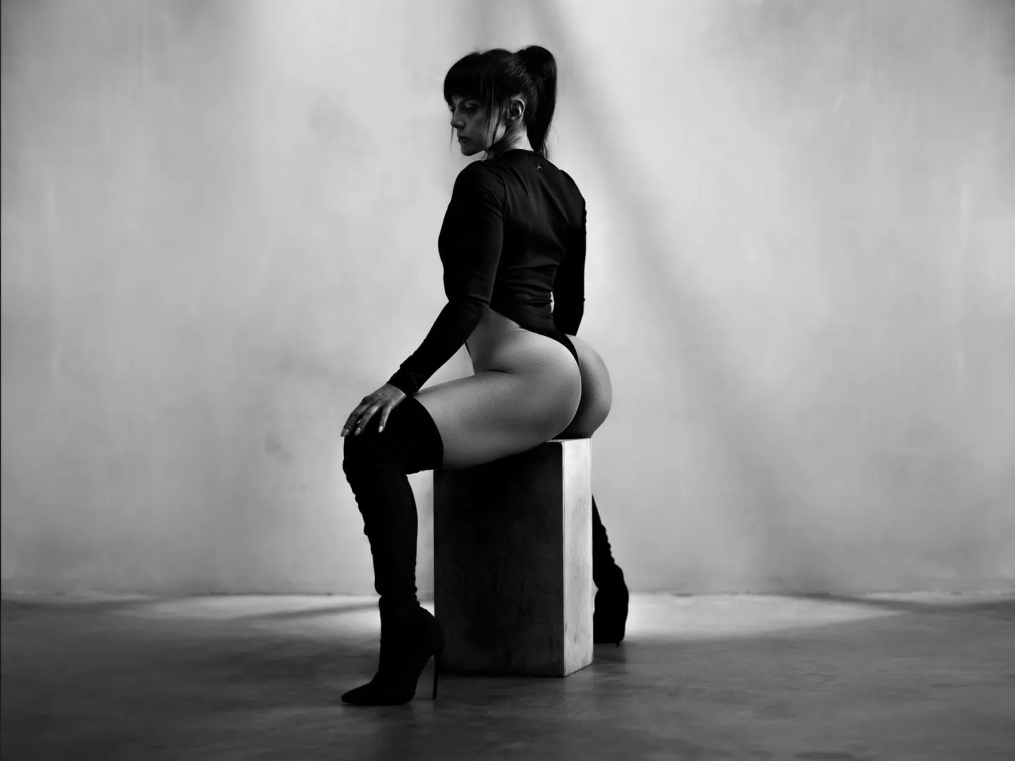 Black and white studio portrait of a woman seated on a wooden plinth, wearing a long-sleeve bodysuit and thigh-high boots, looking over her shoulder