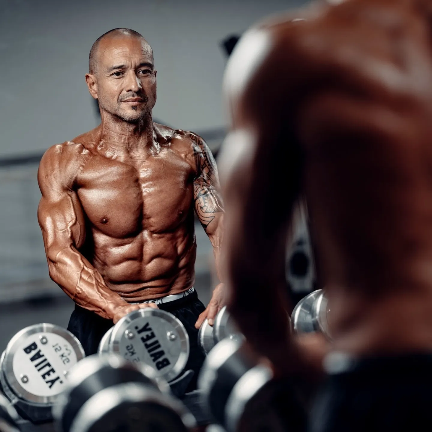 A muscular shirtless male fitness coach leans over a rack of dumbbells in a gym, looking directly at the camera, with a blurred figure in the foreground